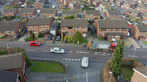 A bin lorry, refuse collection vehicle makes it way up the road as men load recycling bins into the