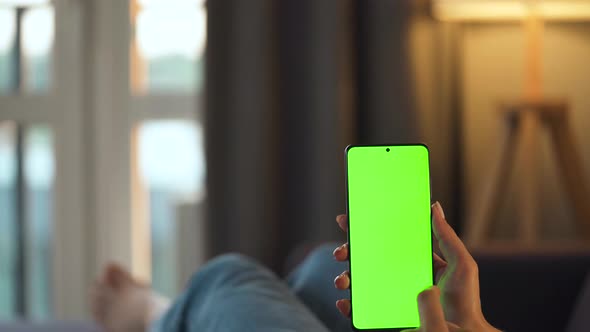 Woman at Home Lying on a Sofa and Using Smartphone with Green Mock-up Screen in Vertical Mode. Girl