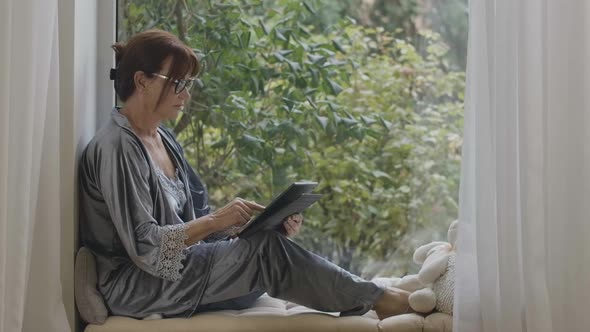 Relaxed Senior Redhead Woman Messaging Online on Tablet Sitting on Windowsill