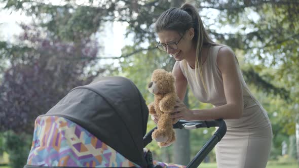 Charming Young Caucasian Woman in Eyeglasses Playing Teddy Bear and Laughing Admiring Infant in Baby