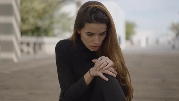Young Shocked Woman Dressed in Black Sits on Street and Looks Down