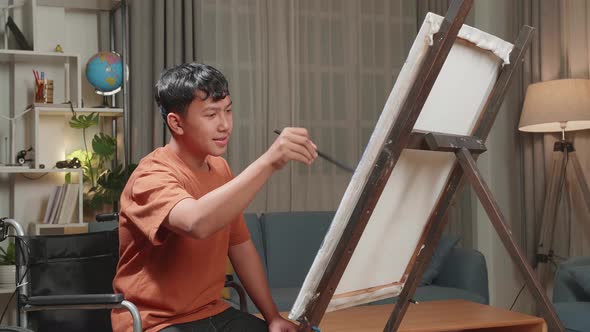 Happy Asian Artist Boy In Wheelchair Holding Paintbrush And Thinking Before Painting On The Canvas