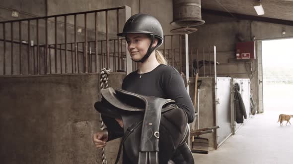 Woman In Riding Gear Carrying Saddle To Horse In Stall Of Stables