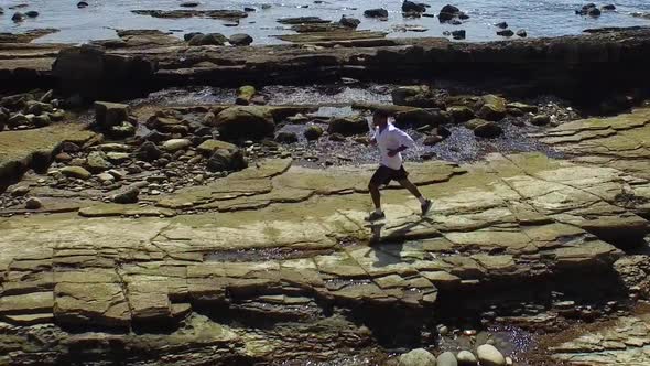 Side view tracking shot of a young man running on a rocky ocean beach shoreline.