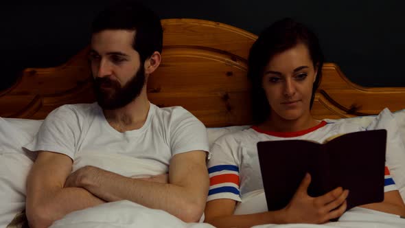 Man getting annoyed while woman reading book on bed