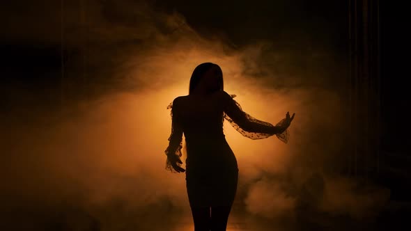 Dark Silhouette of Young Woman Dancing in Dark Smoky Studio Against Background of Yellow Lights