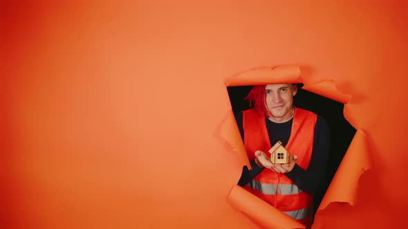 Male Construction Worker in Overalls Holding Small Wooden House and Sticking Out of Hole of Orange