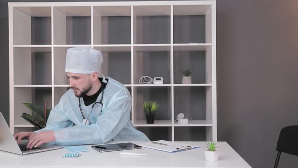 A Doctor Who Works at the Workplace in the Office at the Computer. A Doctor in Medical Clothing Is