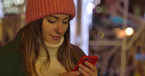 Close-up of Cute Caucasian Girl in Winter Clothes Using Smartphone and Smiling. Young Beautiful