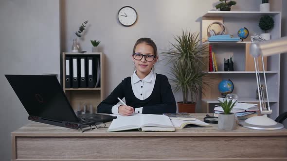 Schoolgirl in Glasses Sitting at the Table Over Her Hometask and Looking at Camera at Home