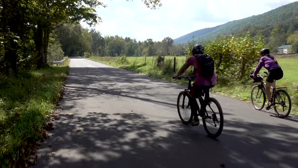 Mother and teenage son biking side by side on a tree covered rural road with a farm in the backgroun