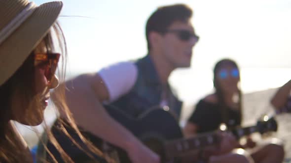 Young Man Playing Guitar Among Group of Friends Sitting on Easychairs on the Beach and Singing on a