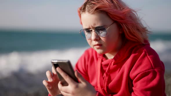 Preteen Girl with Smartphone is Sitting on Seacoast in Cold Windy Weather Watching Social Media