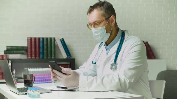 Middleaged Doctor Working with His Smartphone in Front of Laptop in Clinic Office