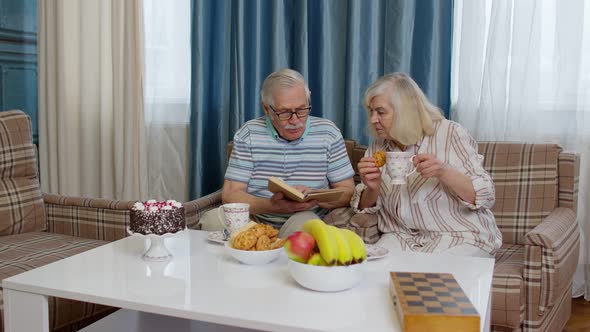 Retired Senior Couple Talking Drinking Tea Reading Book in Modern Living Home Room Lounge Together