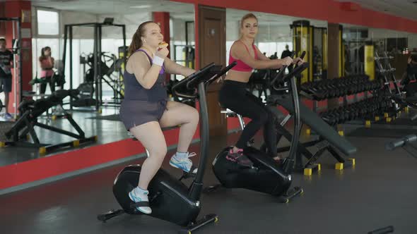 Lazy Corpulent Woman Eating Bun and Riding at Stationary Bike in the Gym