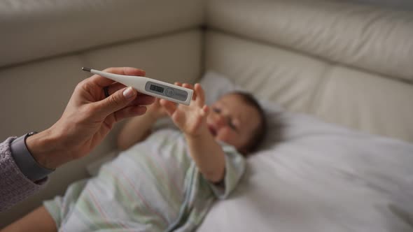 Worried Young Mother Sitting on Sofa Beside Her Sick Son with High Fever