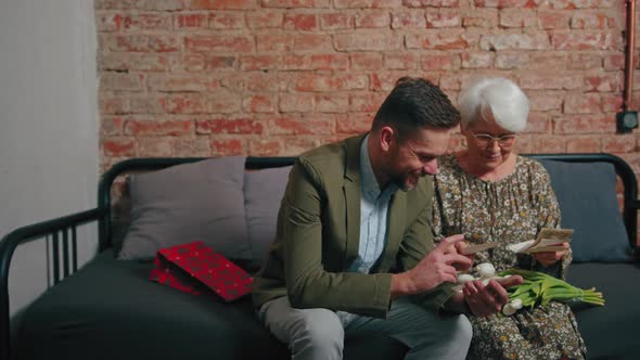 Greyhaired Elderly Pensioner Woman Sitting on a Sofa and Looking at Her Adult Millennial Son's