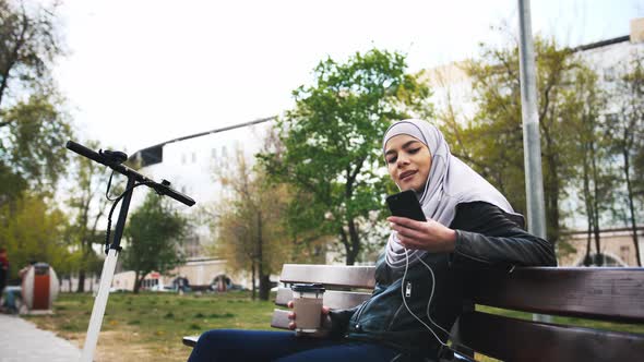 Young Moderm Attractive Muslim Woman Sitting in Park Wearing Hijab and Listening to Music with