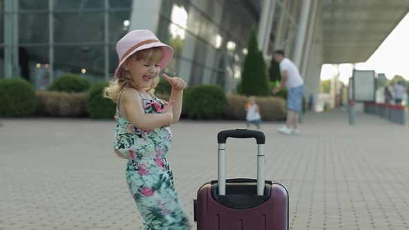 Child Girl Tourist with Suitcase Luggage Bag Near Airport. Kid Dances, Rejoices, Celebrates