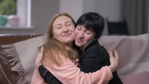 Two Happy Women Hugging Smiling Looking at Camera Sitting in Comfortable Living Room on Cozy Couch