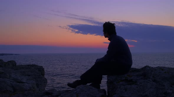 Young Man Waits for the Sunrise at the Sea. Blue hour twilight, gazing at the horizon. Middle Shot