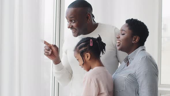 Afro American Happy Family Parents with Little Daughter Looking Out Window Standing at Home