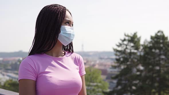 A Young Black Woman in a Face Mask Looks Around - a Cityscape in the Blurry Background