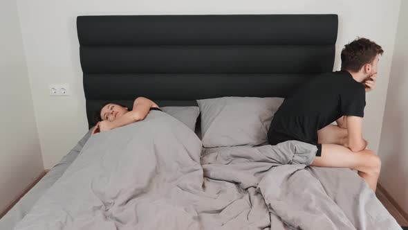 Depressed and stressed man sitting on bed edge, his young wife lies beside him alone. Sex problem