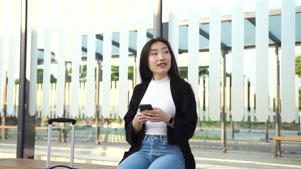 Young Asian Woman in Casual Clothes Sitting on Public Transport Stop and Using Smartphone