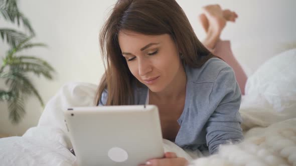 Young Brunette Is Using Gadget, Talking on Phone Lying on Bed Indoors