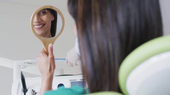 Biracial female patient looking at teeth and smiling in mirror at modern dental clinic