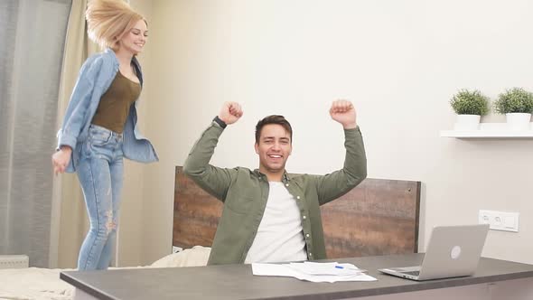 Happy Young Man Sit on Table with Documents While His Girlfriend Jump on Bed