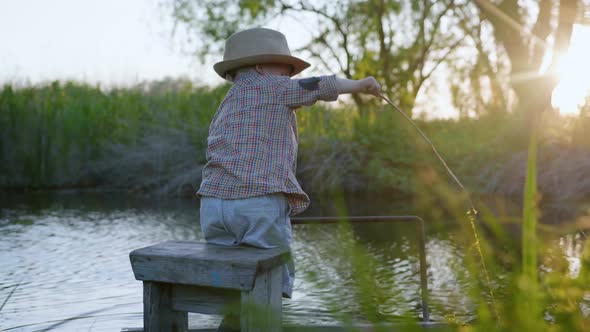 Happy Childhood, a Small Attractive Boy in a Hat Plays with a Stick in the River Having Fun on Pier