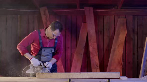 Male Carpenter Cleans Surface of Wooden Beam Using Electric Planer in Workshop