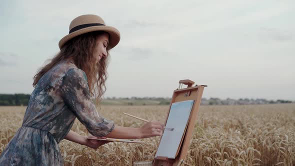 Happy Lady in Hat and Dress Painting Landscape on Canvas Among Wheat Field