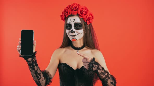 Day of Dead Female with Skull Makeup Raising Up Cellphone Pointing at Its Screen By Forefinger and