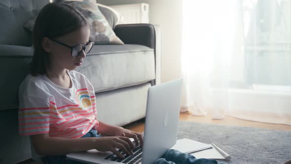 Serious 8 Years Old Girl in Glasses Typing on a Laptop While Sits on a Carpet