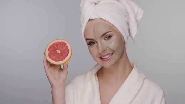 Healthy and Beautiful Woman in Bathrobe and Clay Mask Showing Fresh Grape Fruit Slice