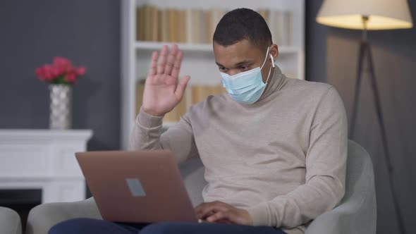 Young Serious African American Man in Coronavirus Face Mask Using Virtual Conference on Laptop As