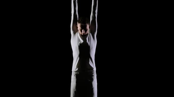Young Handsome Male Acrobat Gymnast in White Clothes on a Black Background Makes Jumps and Shows