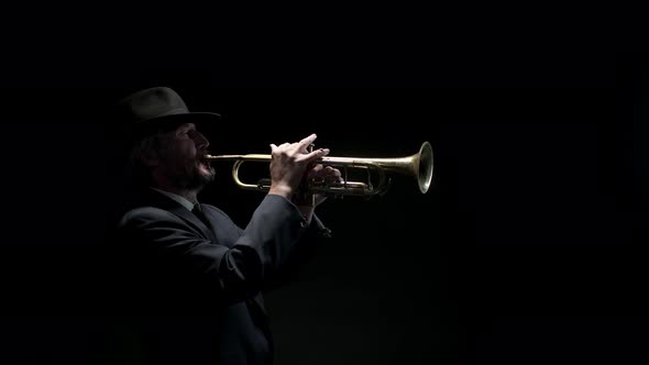 Man in Jacket and Hat Picks Up Trumpet and Plays Music on It at Black Background Side View