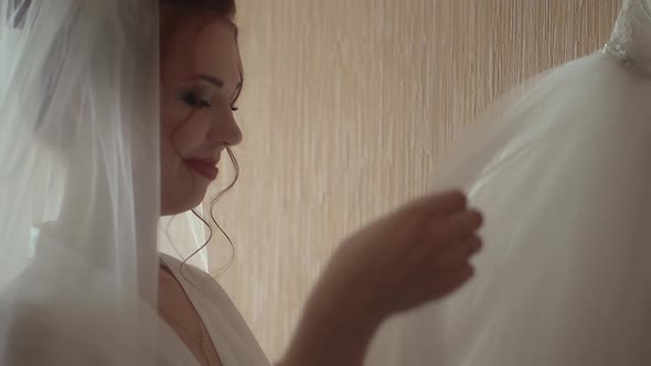 Bride Looks at Wedding Dress Which Hangs on the Wall Looks at It and is Happy Smiles Closeup