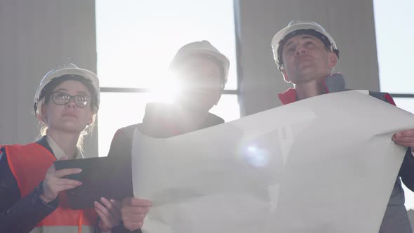 Construction Engineers and Architects in Protective Helmets with Drawings in Hands Discuss