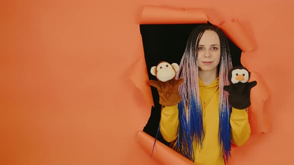 Young Woman with Soft Puppet Toys on Hands Looking Out of Hole of Orange Background