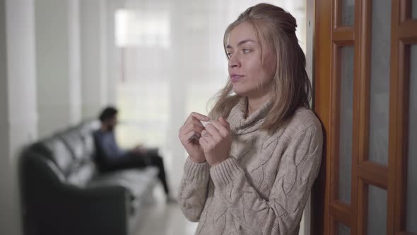 Side View of Blond Caucasian Woman Standing with Pregnancy Test, Looking Back at Husband