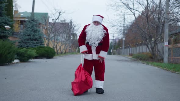 Wide Shot Santa Clause Walking on Suburban Street Having Back Pain Hanging Gift Bag on Shoulder