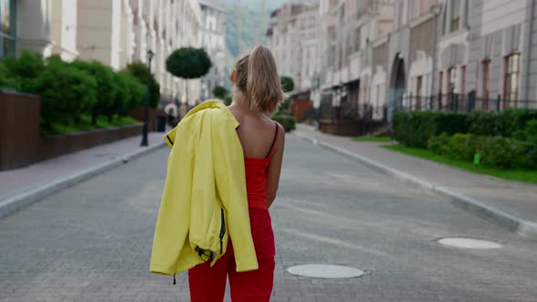 a Woman in Red Clothes Has Thrown a Yellow Jacket Over Shoulder and is Walking Down a City Street