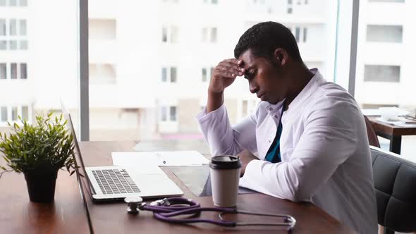 Side View of Tired African American Black Man Doctor Working on Laptop and Drinking Coffee at Desk.