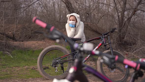 Young Woman in Medical Mask and Gloves Stands Leaning on Bicycle in Countryside